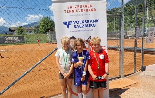 Drei Kinder mit Medaillen stehen vor einer Werbetafel auf einem Tennisplatz in Salzburg.